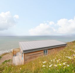 A wooden cabin with a metal roof on a grassy hill overlooking the ocean at Solana in Whitsand Bay