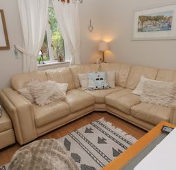 A living room with a cream leather corner sofa and cushions beside a window with white curtains and framed pictures at Albert's Den in Falmouth