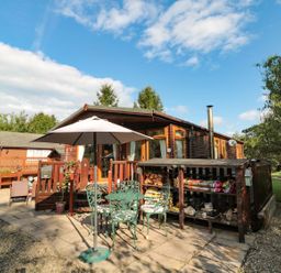 An outdoor patio area with a green metal table and chairs under a white umbrella beside a wooden lodge with a woodpile storage at Evergreen Lodge in Felmoor Holiday Park near Felton