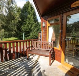 A wooden bench on a deck outside a cabin with glass door and railing at Evergreen Lodge in Felmoor Holiday Park near Felton