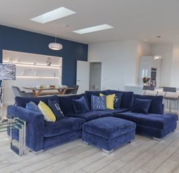 An open living area with a blue sectional sofa and cushions a dining table with chairs and a kitchen with bar stools at The Penthouse in Scarborough