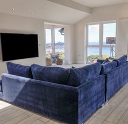 A living room with a blue sofa facing a wall-mounted TV and windows showing a sea view at The Penthouse in Scarborough