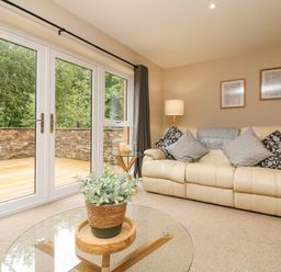 A living room with a cream sofa cushions a glass coffee table with a potted plant a side table with candles and a lamp near large windows at Spicery in Tedburn St Mary
