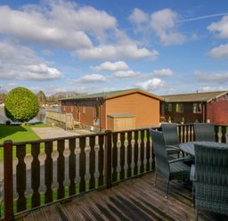 A wooden deck with a round table and four chairs overlooking wooden lodges and a small lake at Lakeside 20 in Warton near Carnforth