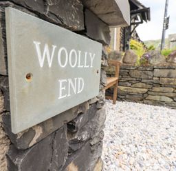 A stone wall with a sign reading Woolly End and a pebble ground area with a wooden bench outside Woolly End Cottage in Ambleside