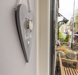 A wooden heart decoration with sheep and text Eve and Me hanging on a wall near a window showing an outdoor bench and stone wall at Woolly End Cottage in Ambleside