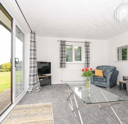 A living room with a glass coffee table with flowers a blue armchair yellow cushions a gray sofa a TV on a stand and sliding glass doors at The Old Forge in Trerulefoot