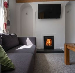A living room with a grey sofa a green cushion a wood burning stove a wall mounted tv and a wooden coffee table at Ayr Farmhouse in St Ives