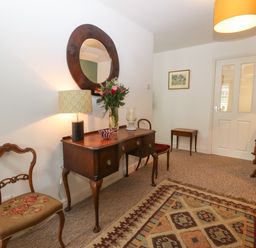 A room with wooden chairs a wooden table with a lamp flowers and a bowl a round mirror on the wall and a patterned rug on the floor at The Bell Apartment in Cemaes Bay