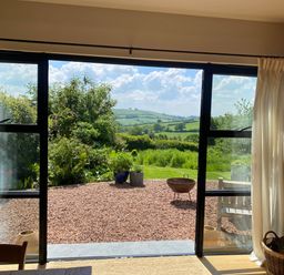 A room with sliding glass doors opening to a gravel patio and garden with hills in the distance at Church Farm Dairy in Cadbury near Thorverton