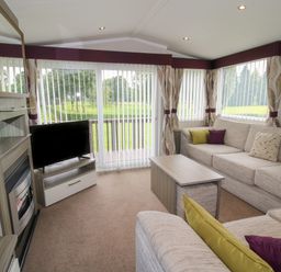 A living room with a beige sectional sofa, coffee table, TV on a stand, fireplace, and large windows with vertical blinds at Swan in Shobdon