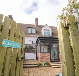 A wooden gate with Coast Cottage sign opening to a brick paved entrance and a house with brick and white walls at Coast Cottage in Bamburgh