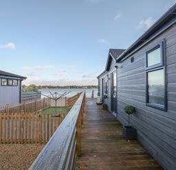 A wooden walkway beside a grey house with potted plants and a fenced garden with drying rack overlooking a lake at Lake View Lodge in Tallington