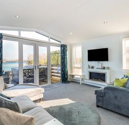 A living room with sofas a wall-mounted TV a fireplace and large glass doors overlooking water at Lake View Lodge in Tallington