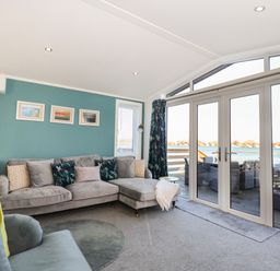 A living room with a grey sectional sofa and armchair facing glass doors with a view of water at Lake View Lodge in Tallington
