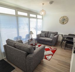 A living room with two grey sofas a small table with a lamp a patterned rug a dining table with chairs a wall clock and a television at 114 Cherry Park in Chapel St Leonards