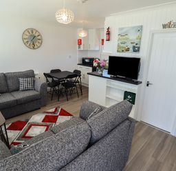 A living room with grey sofas a dining table with four black chairs a TV on a white cabinet and a kitchen area at 114 Cherry Park in Chapel St Leonards