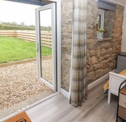 A bedroom with a bed a nightstand with a lamp a small chest of drawers a stone wall and an open glass door leading to a garden at The Nest in Amroth