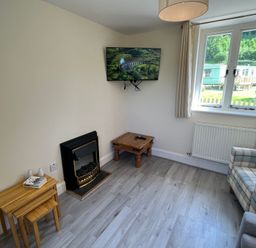A small living room with a mounted television above a fireplace two nested wooden tables a wooden side table with remotes a plaid armchair next to a window with curtains at Railway Station Cottage in New Radnor near Kington