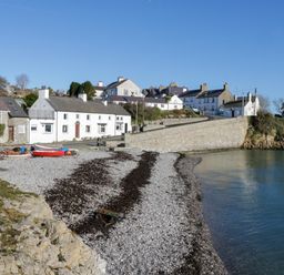 A pebble beach with boats near a stone seawall and houses in a coastal village at 3 Moranedd in Moelfre