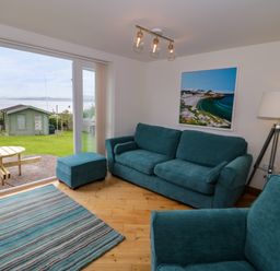 A living room with blue sofas and a rug next to a glass door overlooking a garden with a shed at 3 Moranedd in Moelfre