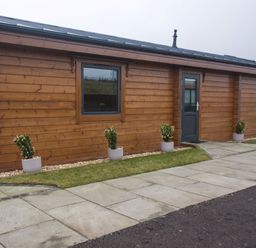 A wooden cabin exterior with potted plants along the wall and a paved walkway at Height End Farm Log Cabin in Rossendale near Bacup
