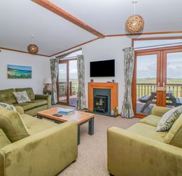 A living room with green sofas a wooden coffee table a wall-mounted TV and glass doors opening to a balcony at Three Views Lodge in Whitsand Bay Fort Holiday Village