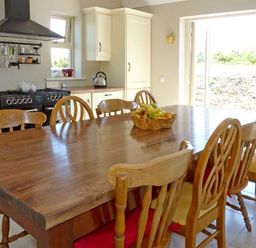 A kitchen with a wooden dining table and chairs a fruit bowl on the table and a black stove with cabinets at Bluebell Cottage in Spanish Point County Clare