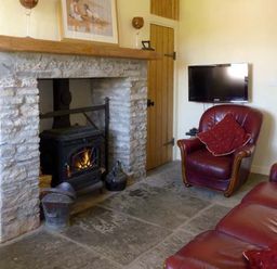 A living room with a stone fireplace wood burning stove red leather armchair and sofa and a wall mounted television at Bluebell Cottage in Spanish Point County Clare