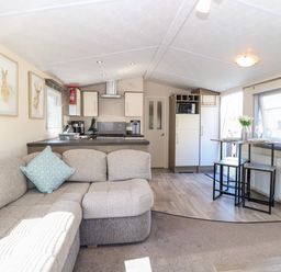 A living room with a beige sectional sofa a small dining table with two stools and a kitchen area at Hazell Thorne Hideaway in Fritton