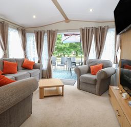 A living room with gray sofas and orange cushions a wooden coffee table a wall mounted television and an outdoor seating area at Stanleys Lodge Foxburrow Hang in Belton Norfolk
