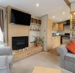 A living room with a wall-mounted TV and fireplace two grey sofas with orange cushions and an open kitchen at Stanleys Lodge Foxburrow Hang in Belton Norfolk
