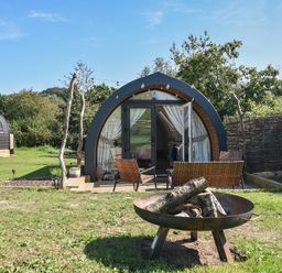 A grassy outdoor area with a fire pit and two woven chairs in front of a pod-shaped cabin with glass doors