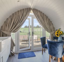 A dining area with blue chairs and sunflowers on a glass table near large windows with curtains at Cedar in Wootton Fitzpaine near Charmouth