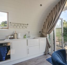 A kitchen with white cabinets a sink an oven and a kettle next to an open glass door leading to a patio at Cedar in Wootton Fitzpaine near Charmouth