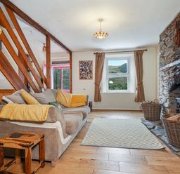A living room with a sofa yellow cushions a wooden side table a stone fireplace and windows with curtains at Delfryn Goginan in Goginan near Aberystwyth