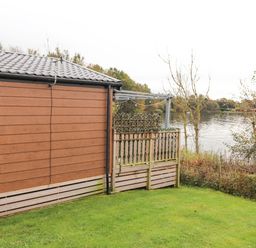 A brown wooden house next to a wooden fence with a lake and trees in the background at Pine Lake View in Coniston View 3 Carnforth