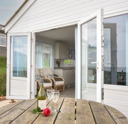 An open doorway leading to a room with armchairs and a kitchen area with flowers on the counter a wooden table with a bottle of wine two glasses and a red rose outside at Sealark in Eype