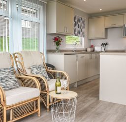 A kitchen with cabinets a microwave a sink and two wicker armchairs with cushions and a small table with a bottle and glasses at Sealark in Eype