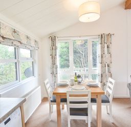 A dining area with a wooden table set for four near windows with floral curtains and a grey sofa at The Hideaway in Eype