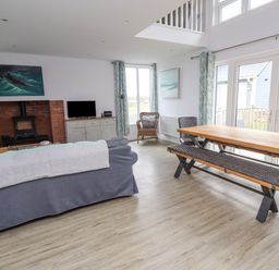 A living room with a gray sofa a wooden dining table with bench and chairs a fireplace and large windows at Turning Tide in Beadnell