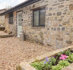 A stone building with a white door and a window and a flower bed with pink and purple flowers in front at The Stable in Amroth