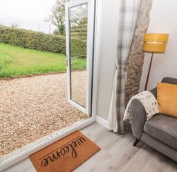 An open glass door leading to a gravel outdoor area with grass and a hedge inside a room with a grey sofa yellow cushion and floor lamp at The Stable in Amroth