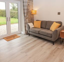 A sitting room with a grey sofa and yellow cushions next to a small wooden side table and glass doors with curtains at The Stable in Amroth