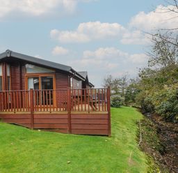A wooden cabin with a deck and railing on a grassy lawn next to a small creek at Haven Lodge in Bossiney Bay near Tintagel