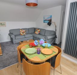 A living room with a gray corner sofa and a wooden dining table with four plates and flowers at The Loft @ Wisteria Cottage in Saundersfoot