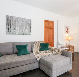 A living room with a gray sectional sofa green pillows a wooden door a white chair a lamp and wall art at Watersong in Coniston