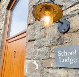 A stone wall with a wooden door a light fixture and a sign that reads School Lodge in Llandeilo