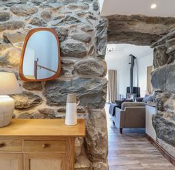 A wooden cabinet with a lamp and vase beside a stone wall opening leading to a living room with a stove at Gaerau Bach in Llanaber
