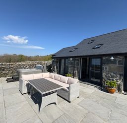 An outdoor patio with a stone floor a rattan sofa set and a hot tub next to a stone building at Gaerau Bach in Llanaber